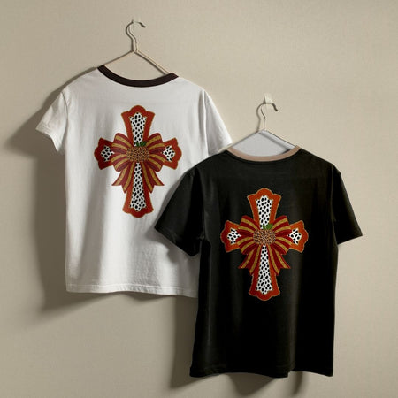 Two short-sleeve T-shirts hanging on hangers against a plain beige wall. The back of one white shirt and one black shirt both feature a large decorative cross graphic in rust red, black, white, and a small green accent.