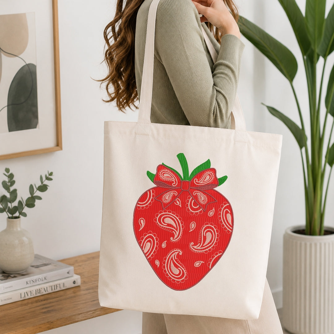 A woman wearing a sage green ribbed long-sleeve top carries a cream canvas tote bag featuring a large red strawberry graphic with white paisley patterns and green leaves.
