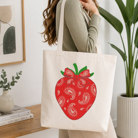 A woman wearing a sage green ribbed long-sleeve top carries a cream canvas tote bag featuring a large red strawberry graphic with white paisley patterns and green leaves.