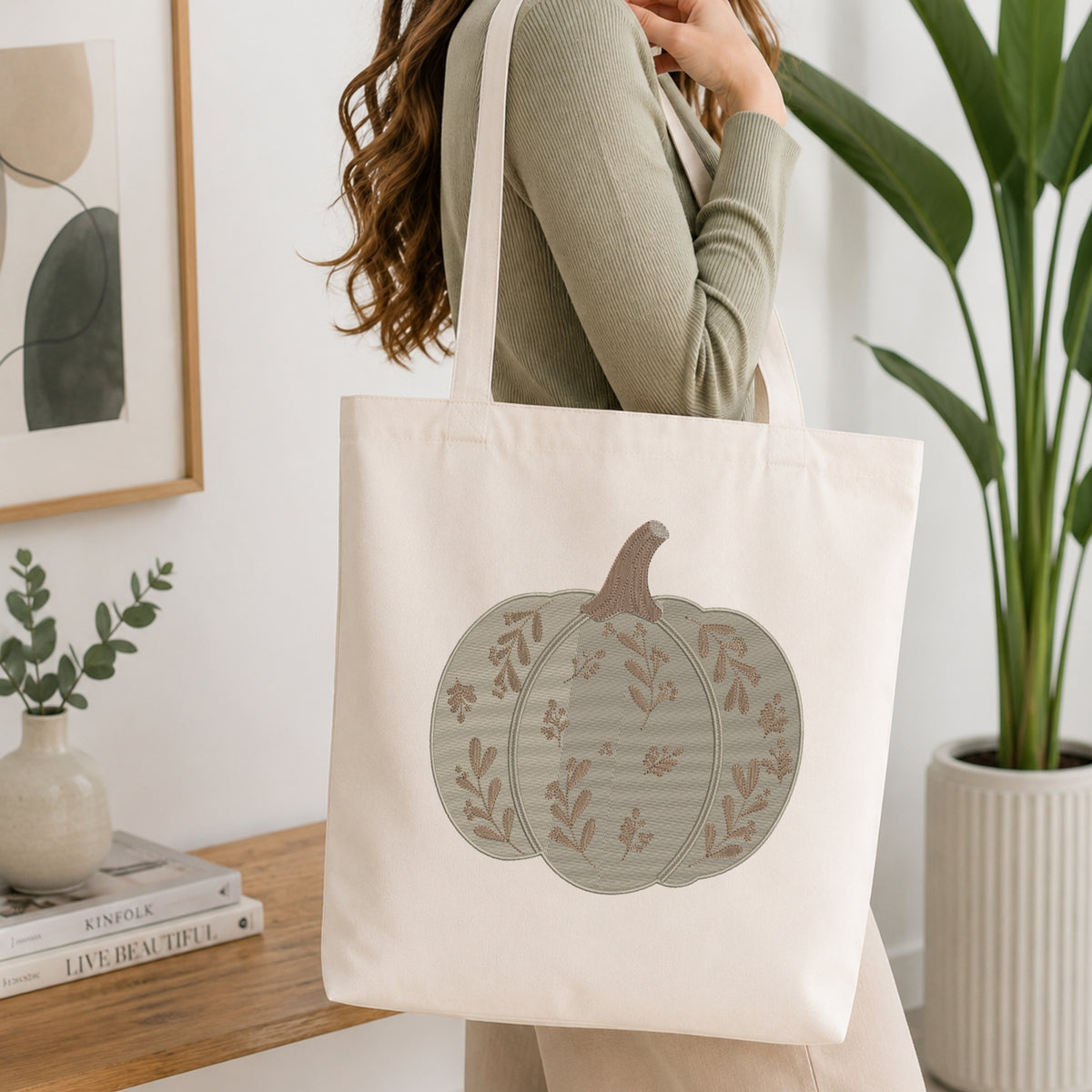 A woman wearing a sage green ribbed sweater carries a cream canvas tote bag with a large pumpkin graphic printed on the front. The background is a bright, modern room with a wooden table, books, a framed print, and a potted plant.