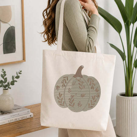 A woman wearing a sage green ribbed sweater carries a cream canvas tote bag with a large pumpkin graphic printed on the front. The background is a bright, modern room with a wooden table, books, a framed print, and a potted plant.