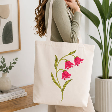A woman wearing a sage green ribbed sweater carries a cream canvas tote bag with embroidered pink bell-shaped flowers and green leaves, set in a bright, modern room with a wooden table and potted plants.