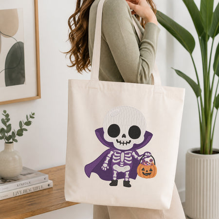 A woman wearing a sage green ribbed sweater carries a cream canvas tote bag with a cartoon skeleton dressed in a purple cape holding an orange pumpkin bucket. The scene is set in a bright, modern room with a wooden table, books, a framed print, and a potted plant.