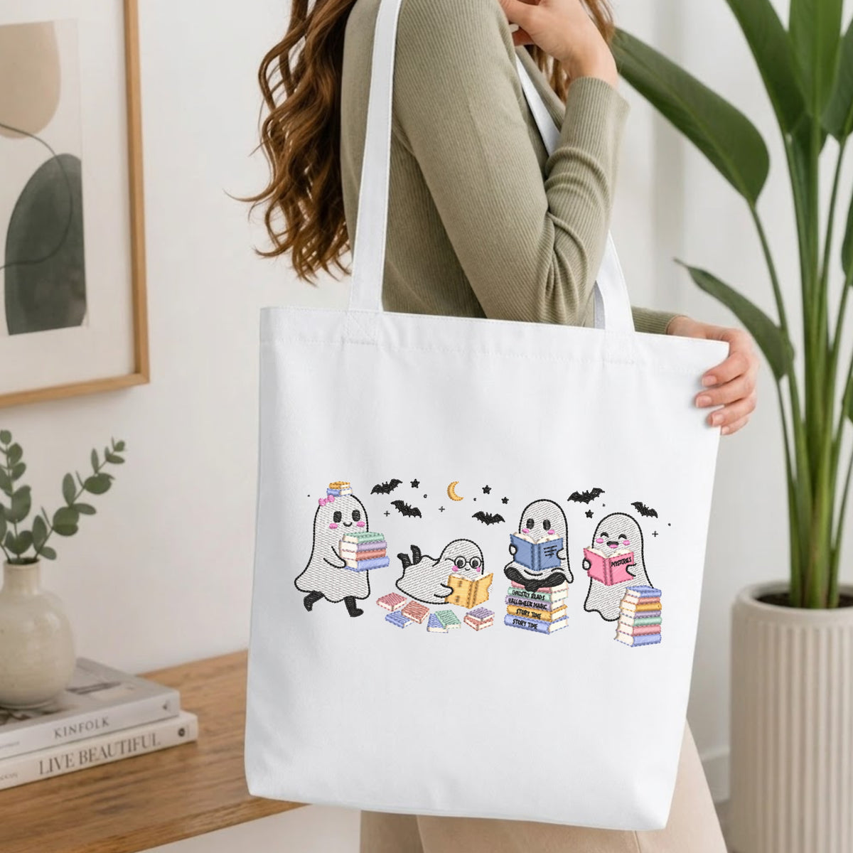 A woman wearing a sage green ribbed long-sleeve top carries a white canvas tote bag. The bag features a cute graphic of four cartoon ghosts reading books, decorated with small black bats and a crescent moon.