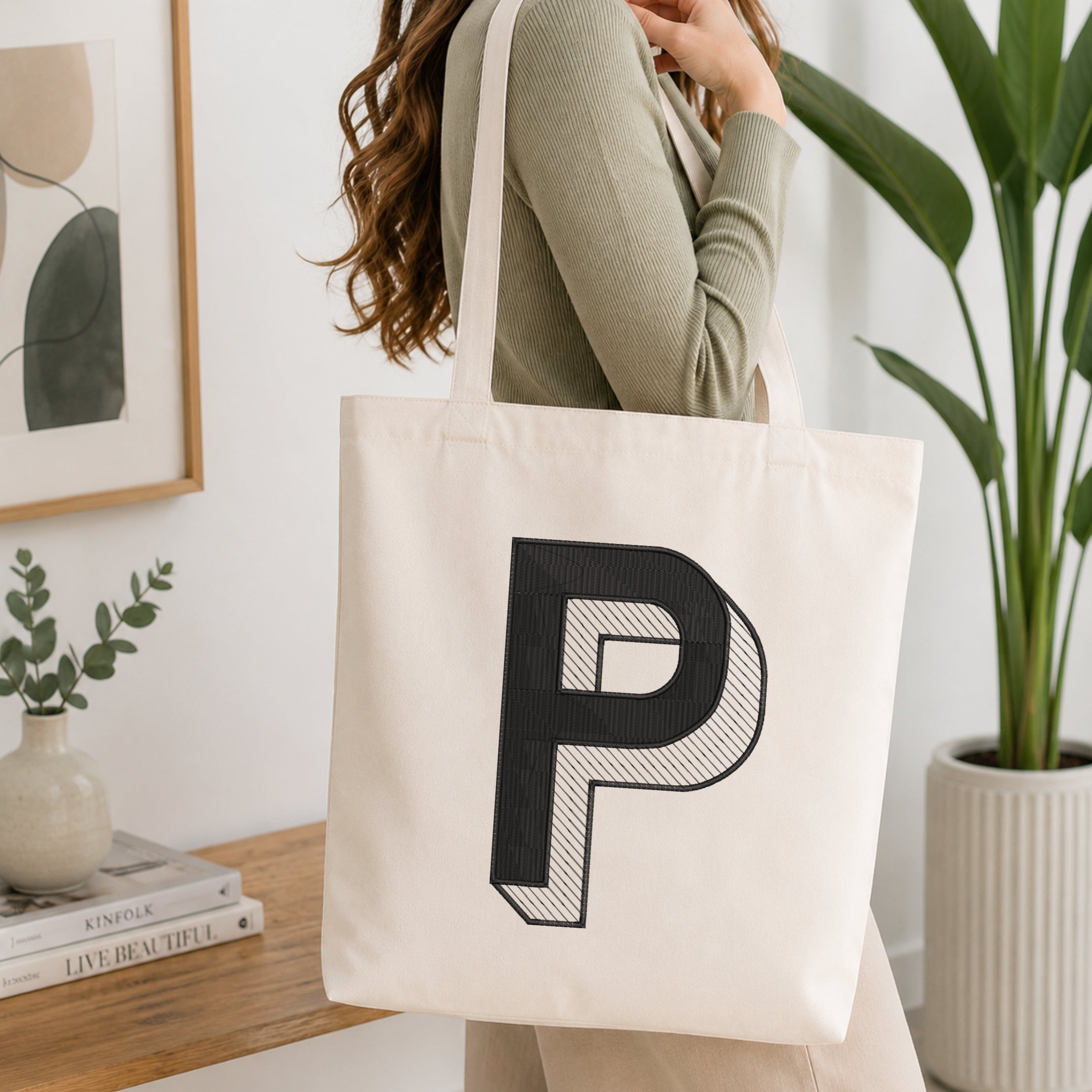A woman wearing a sage green ribbed long-sleeve top carries a cream canvas tote bag with a large black embroidered letter 'P' on the front, set in a bright, modern room with a wooden table, framed art, and a potted plant.