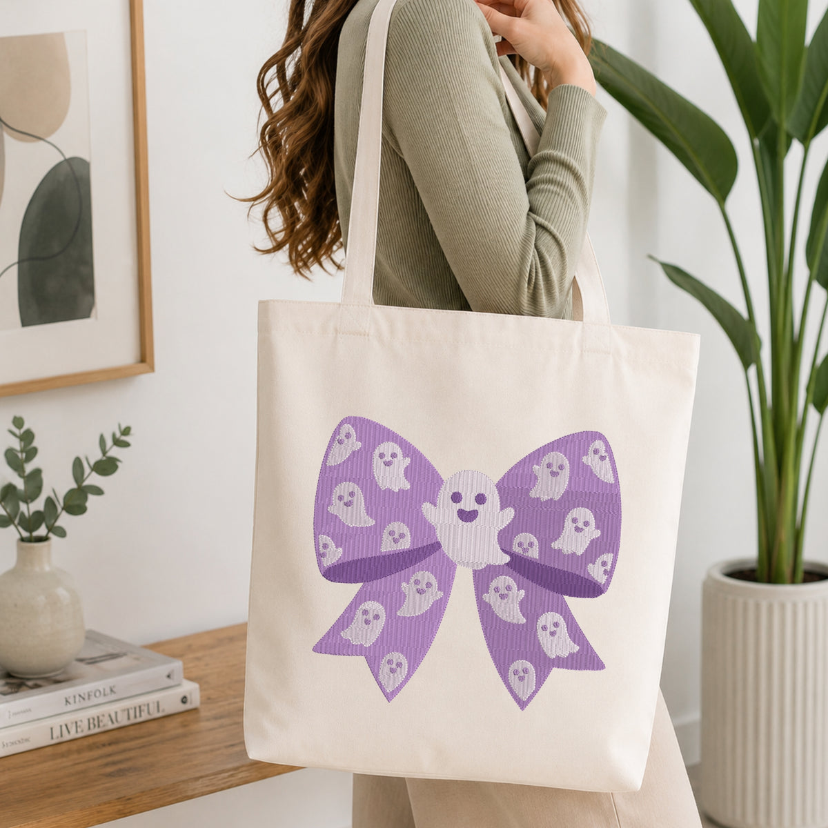 A person wearing a green long-sleeve top and carrying a natural canvas tote bag printed with the purple ghost bow machine embroidery graphic.