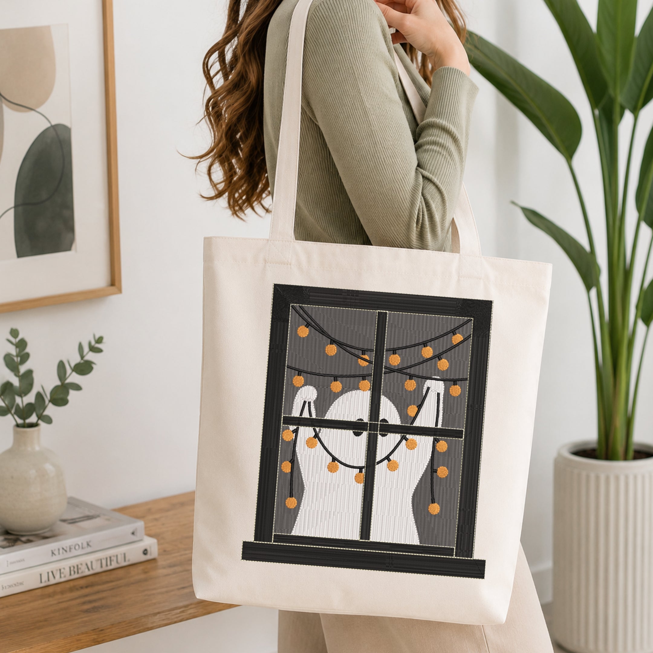 A woman wearing a sage green ribbed sweater carries a cream canvas tote bag with a black-framed window graphic showing a white ghost and orange string lights, set against a bright, modern interior background with a potted plant and books.