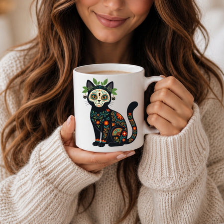 A woman in a cream knit sweater holds a white ceramic mug featuring a colorful sugar-skull cat illustration with green leaves and orange floral details.