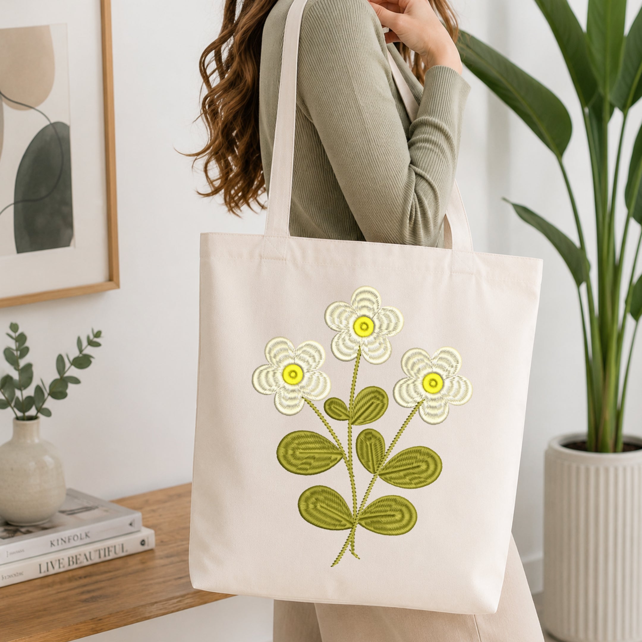 A woman wearing a sage green ribbed sweater carries a cream canvas tote bag with embroidered flowers. The bag features three cream-colored flowers with yellow centers and olive green leaves.