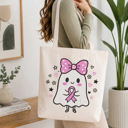 A person wearing a green long-sleeve top and carrying a natural canvas tote bag printed with the ghost holding a pink ribbon machine embroidery design.