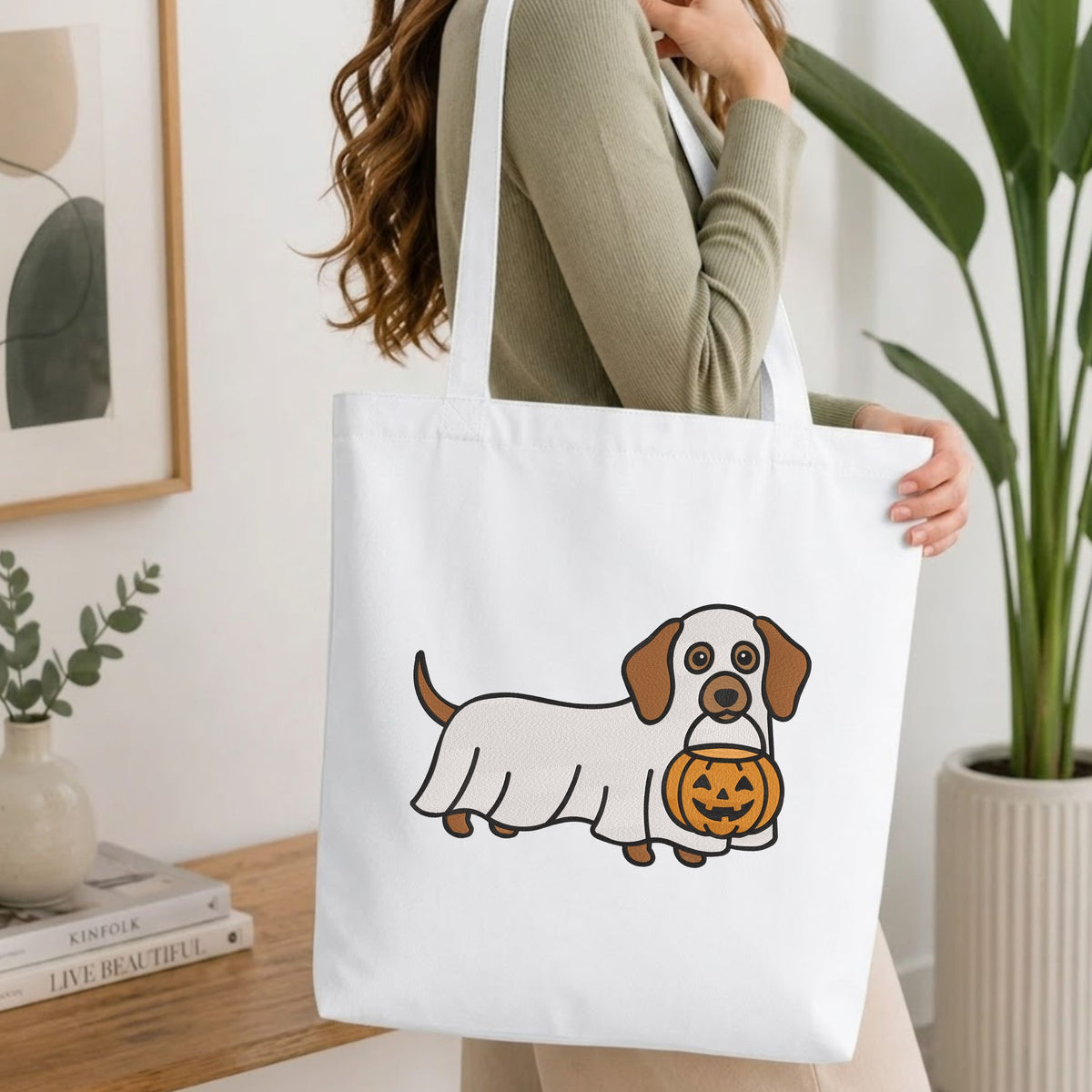 Woman carrying a white natural canvas tote bag featuring a ghost dachshund machine embroidery design in a bright, modern interior setting
