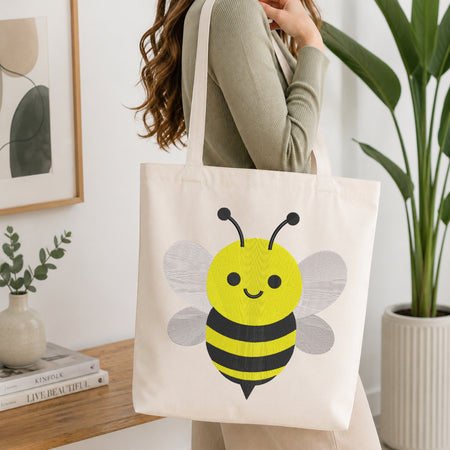 A woman wearing a sage green ribbed sweater carries a cream canvas tote bag featuring a cute yellow and black striped bumblebee graphic. The scene is set in a bright, modern room with a wooden side table, a framed print, a small vase with greenery, and a large potted plant.
