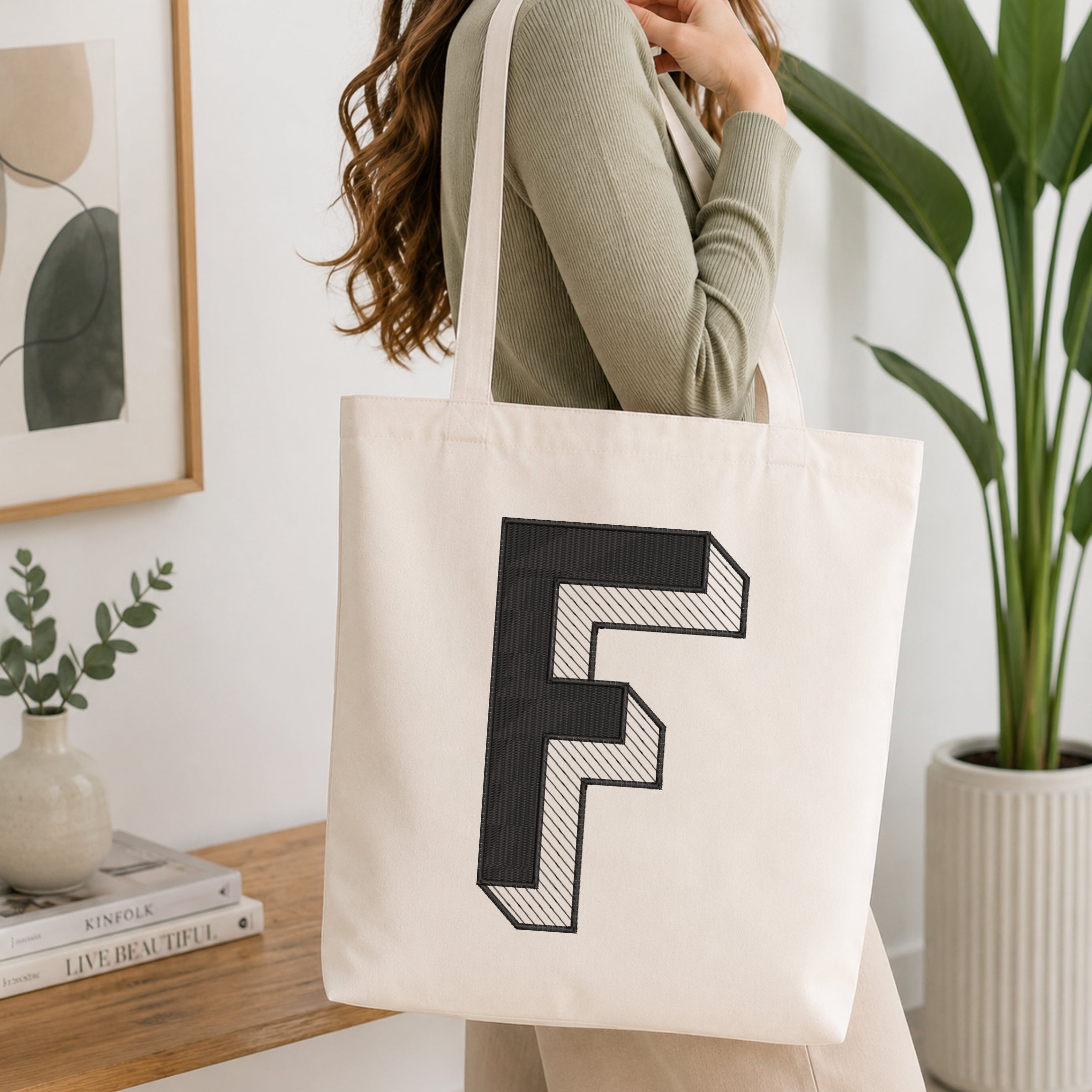 A woman wearing a sage green ribbed long-sleeve top carries a large off-white canvas tote bag with a bold black geometric letter 'F' printed on the front, set in a bright, modern room with a wooden table, framed art, and a potted plant.