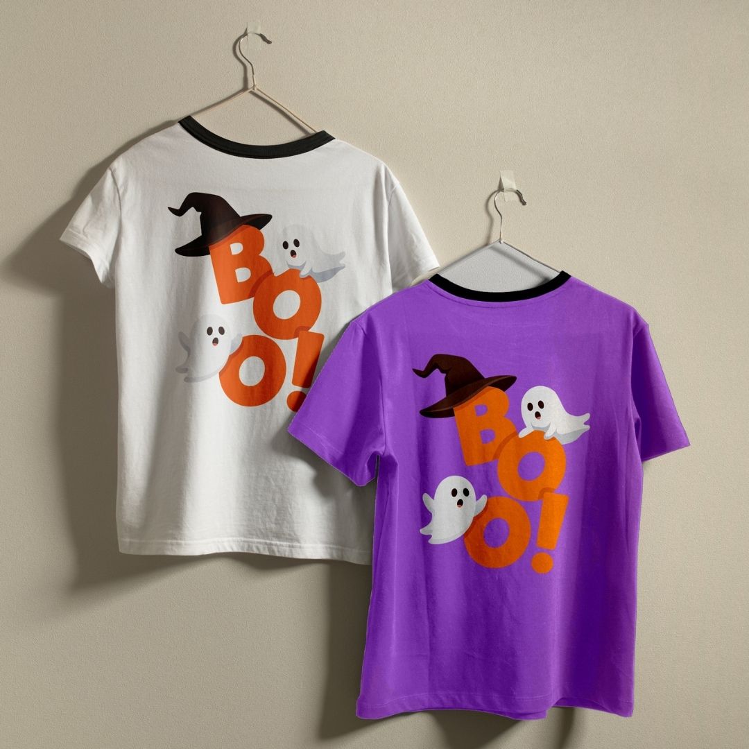 Two t-shirts hanging on a wall—one white and one purple—both featuring the orange "BOO!" ghost and witch hat back print design.