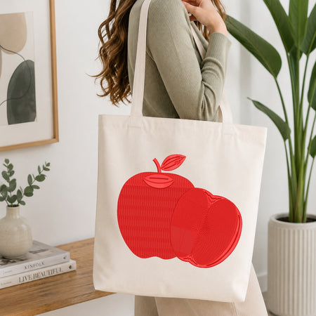 A person wearing a ribbed sage green top carries a cream canvas tote bag with a large red apple graphic on the front, set in a bright, modern room with a wooden table, books, and a potted plant.