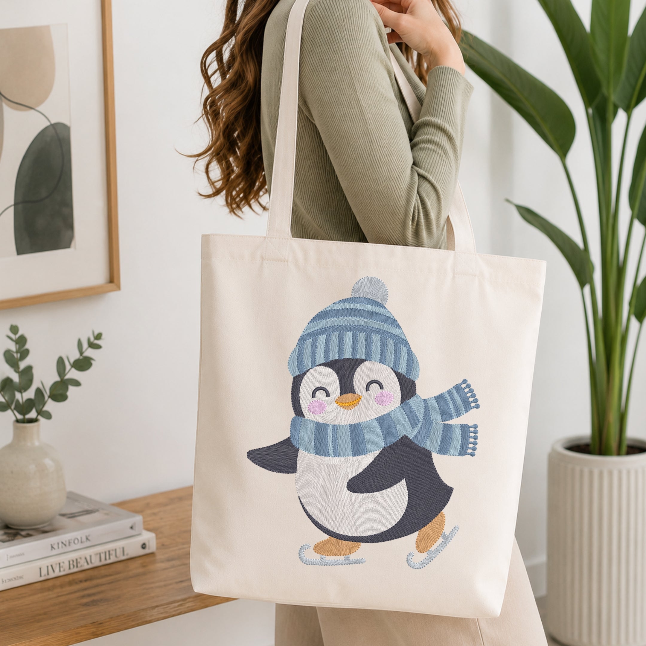 A woman holds a cream canvas tote bag featuring a cute embroidered penguin wearing a blue striped knit hat and matching scarf, set against a bright, modern interior background with a wooden table and potted plants.