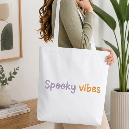 Woman carrying a white natural canvas tote bag featuring a spooky vibes machine embroidery design in a bright, modern interior setting