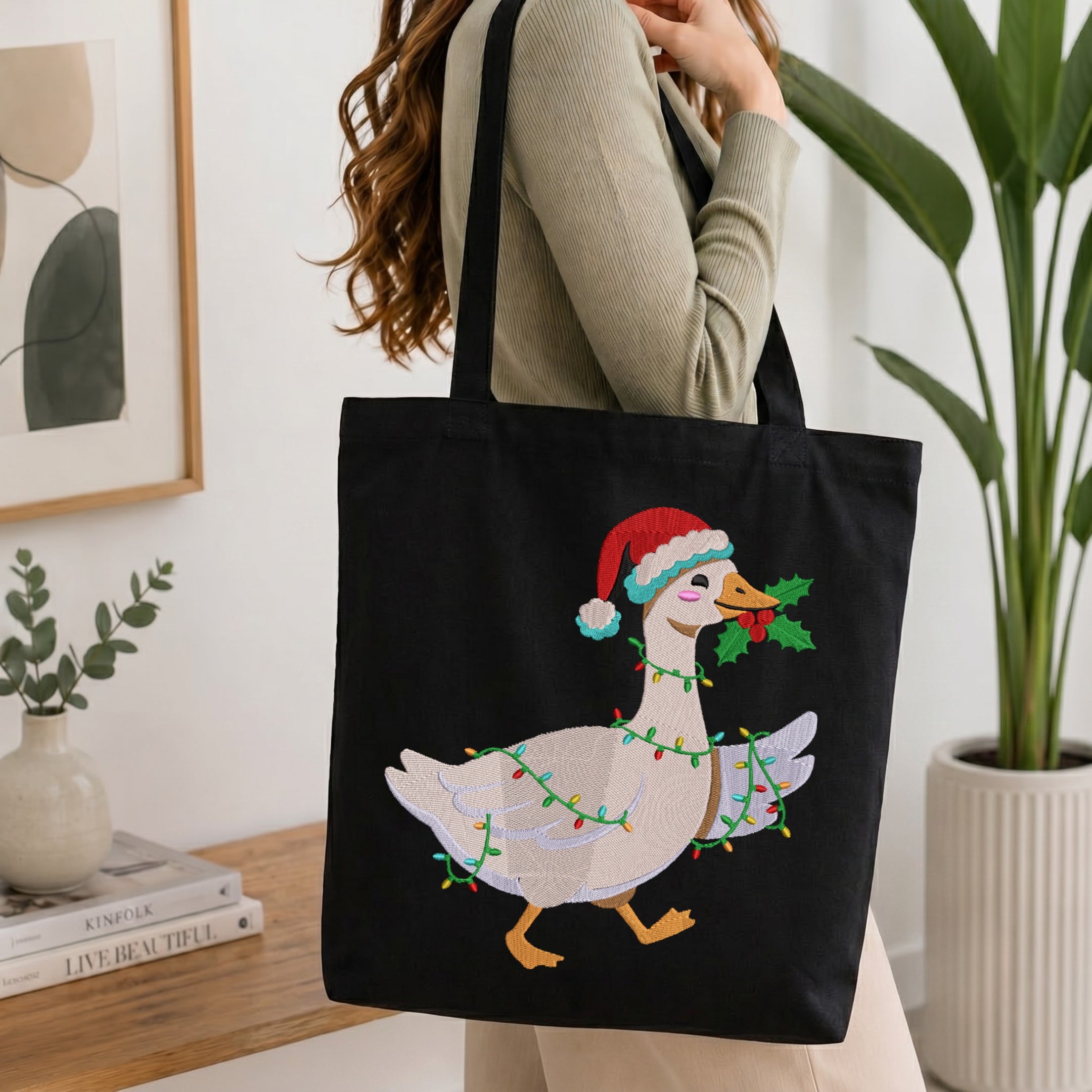 A person carrying a black canvas tote bag embroidered with a festive Christmas goose wearing a Santa hat, wrapped in colorful string lights, and holding a sprig of holly in its beak.