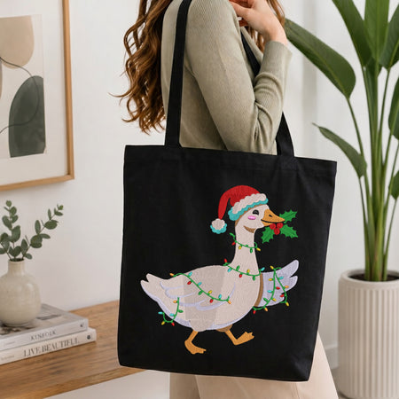 A person carrying a black canvas tote bag embroidered with a festive Christmas goose wearing a Santa hat, wrapped in colorful string lights, and holding a sprig of holly in its beak.