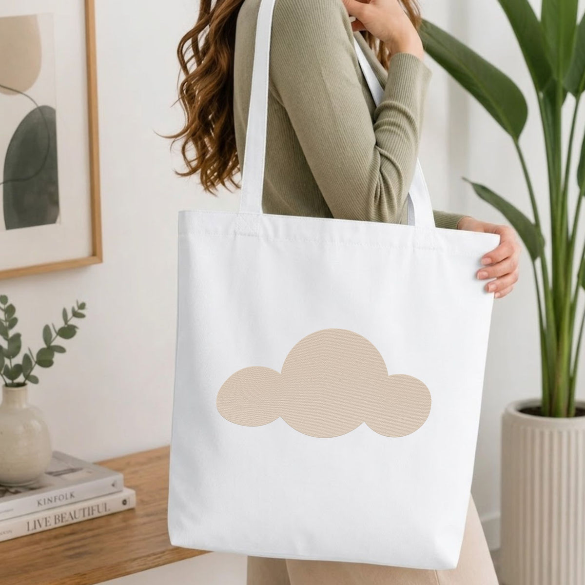 Woman carrying a white natural canvas tote bag featuring a minimalist cloud machine embroidery design in a bright, modern interior setting