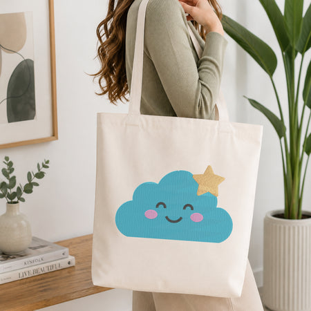A woman wearing a sage green ribbed long-sleeve top carries a cream canvas tote bag with a cute blue cloud and gold glitter star graphic, set in a bright, modern room with a wooden table and potted plants.