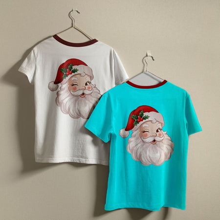 Two short-sleeve Christmas-themed t-shirts hanging on hangers against a plain beige wall. The left shirt is white with a dark red collar and features a large graphic of Santa's face on the back.