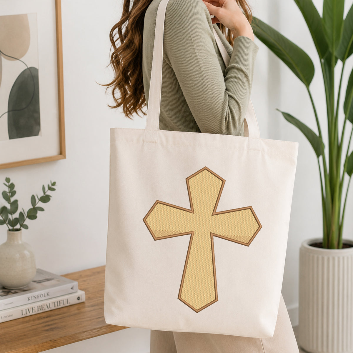 A woman wearing a sage green ribbed top carries a cream canvas tote bag with a large gold cross design outlined in brown, shown in a bright, modern room with a wooden table, books, and a potted plant.