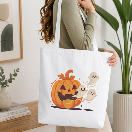 Woman carrying a white natural canvas tote bag featuring an embroidered Halloween pumpkin and ghosts design in a bright, modern interior setting.