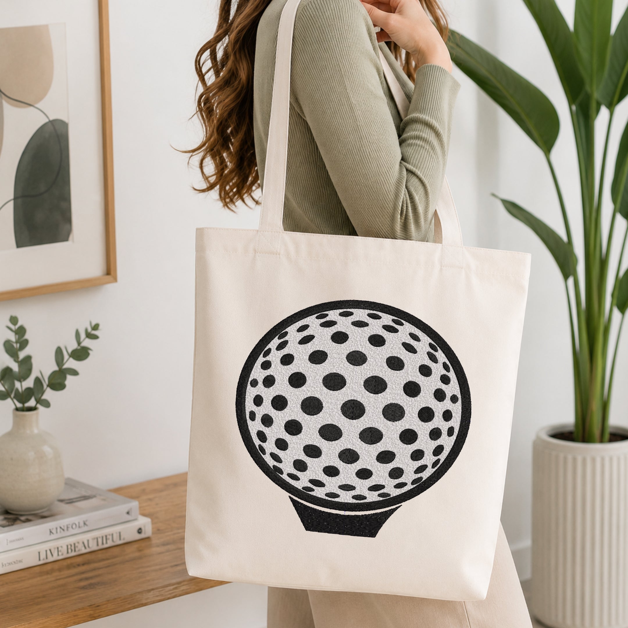 A woman wearing a sage green ribbed sweater carries a cream canvas tote bag with a black graphic of a golf ball on the front, set in a bright, modern room with a wooden table, books, and a potted plant.