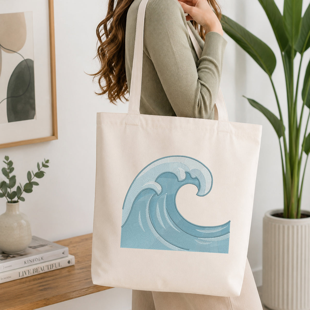 A woman wearing a sage green ribbed long-sleeve top carries a cream canvas tote bag with a blue water wave graphic printed on the front, set in a bright, modern room with a wooden table, framed art, and a potted plant.