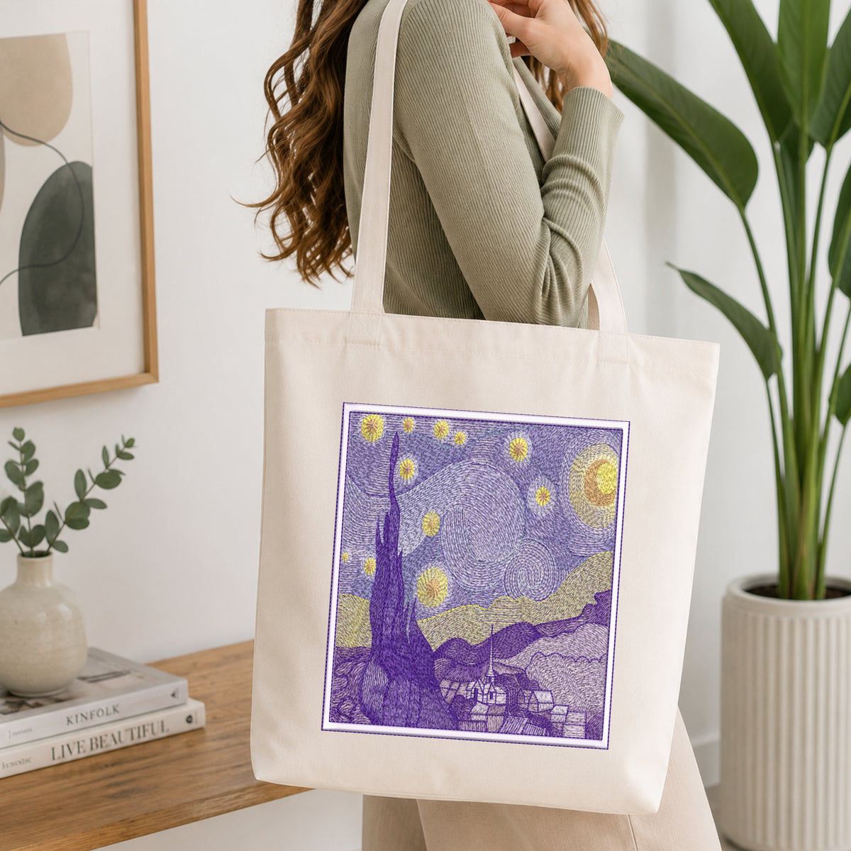 A woman wearing a ribbed sage green top carries a cream canvas tote bag with a purple and yellow Starry Night style graphic print on the front, set in a bright, modern room with a wooden table, books, and a potted plant.