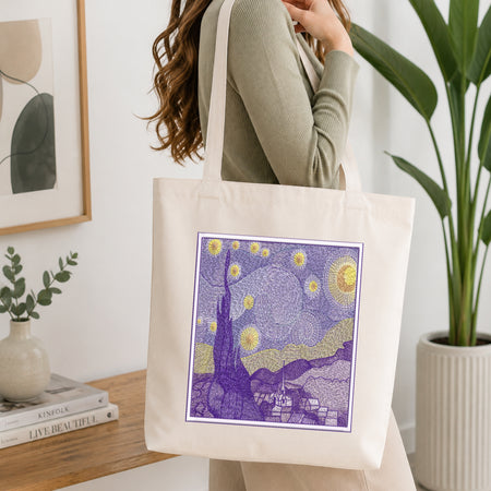 A woman wearing a ribbed sage green top carries a cream canvas tote bag with a purple and yellow Starry Night style graphic print on the front, set in a bright, modern room with a wooden table, books, and a potted plant.