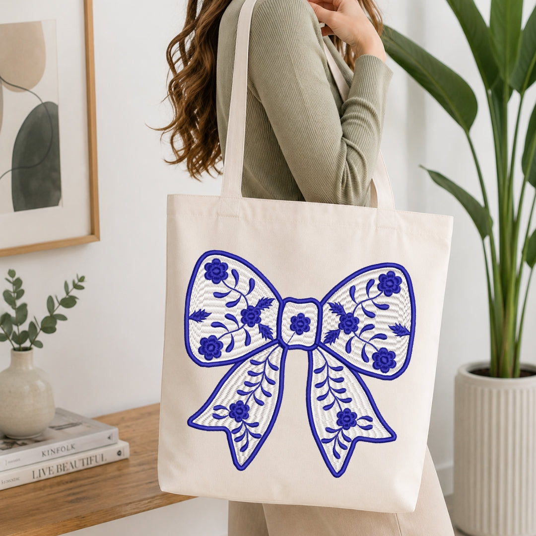 A person wearing a sage green ribbed sweater carries a cream canvas tote bag decorated with a large blue floral bow design, set in a bright, modern room with a wooden table and potted plants.