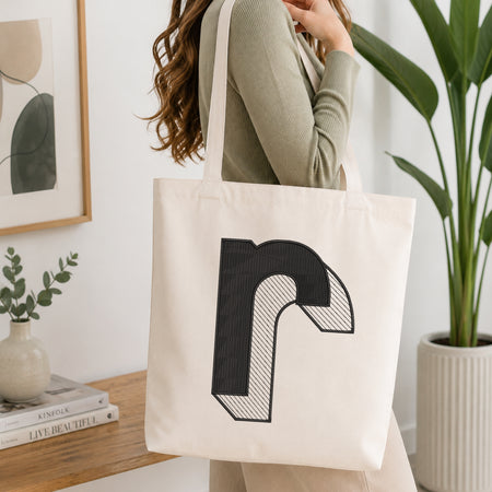 A woman wearing a sage green ribbed long-sleeve top carries a large off-white canvas tote bag with a bold black graphic letter 'R' printed on the front, set in a bright, modern room with a wooden table, books, and a potted plant.
