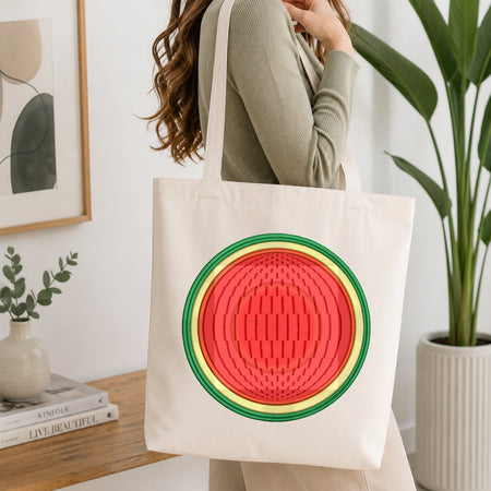 A person wearing a ribbed sage green top carries a cream canvas tote bag featuring a large, colorful watermelon graphic on the front. The setting is a bright, modern room with a wooden table, books, a small vase, and a potted green plant in the background.