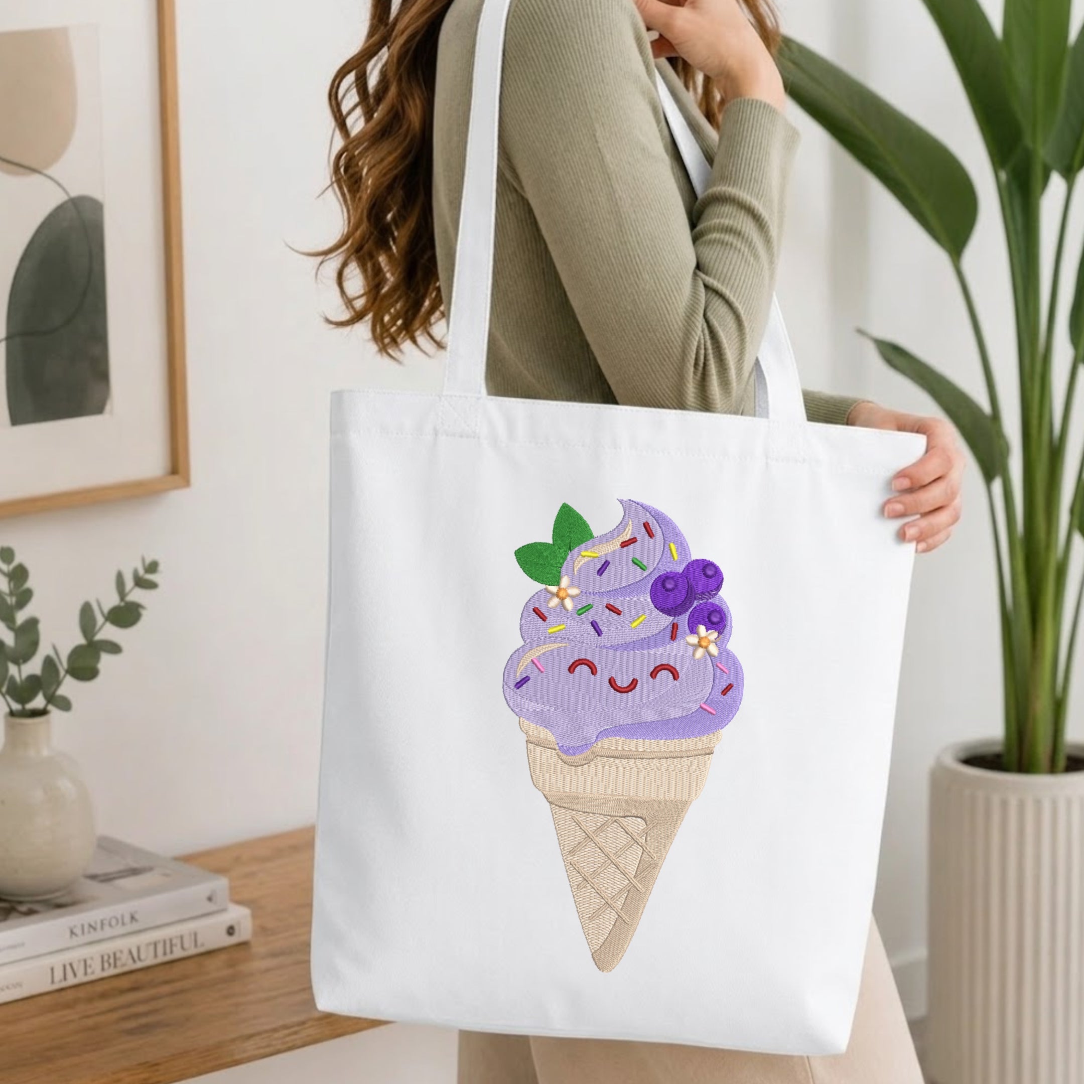 A woman in a sage green ribbed sweater carries a white canvas tote bag featuring a colorful embroidered ice cream cone design with purple swirls, blueberries, and sprinkles, set against a bright, modern indoor background.