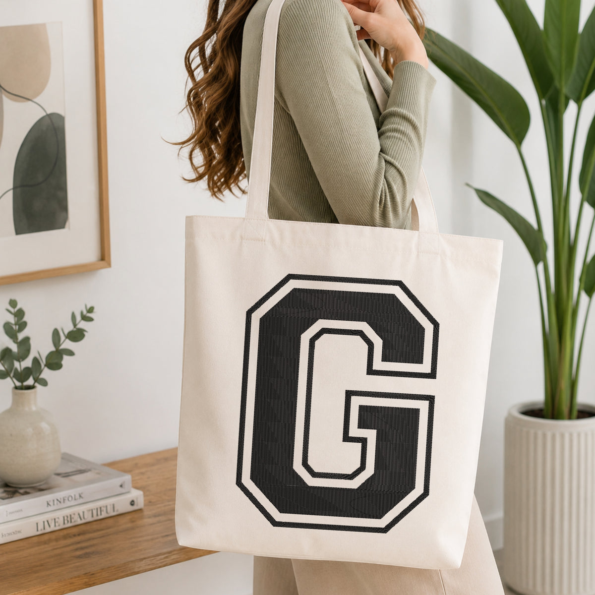 A woman wearing a sage green ribbed long-sleeve top carries a large off-white canvas tote bag with a bold black collegiate letter 'G' printed on the front, set in a bright, modern room with a wooden console table and a potted plant.