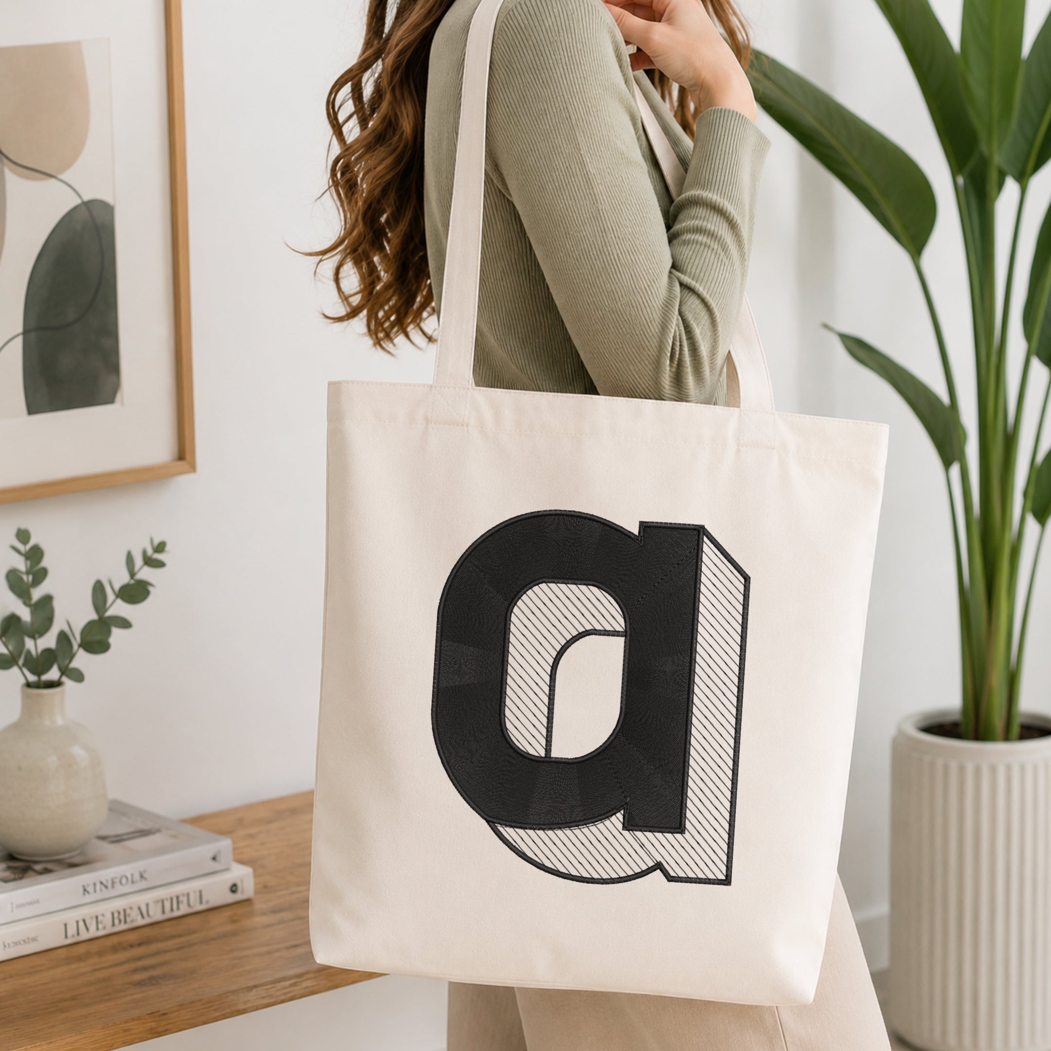 A woman wearing a sage green ribbed long-sleeve top carries a cream canvas tote bag with a large black stylized letter 'a' graphic on the front, set in a bright, modern room with a wooden table, books, and a potted plant.