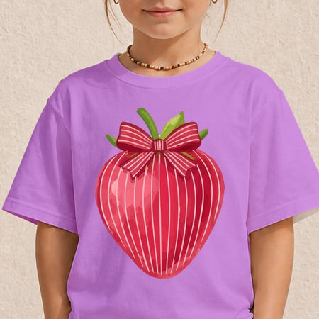 A child wearing a lavender purple t-shirt with a large graphic of a red striped strawberry wearing a pink striped bow on the front, set against a textured beige wall.