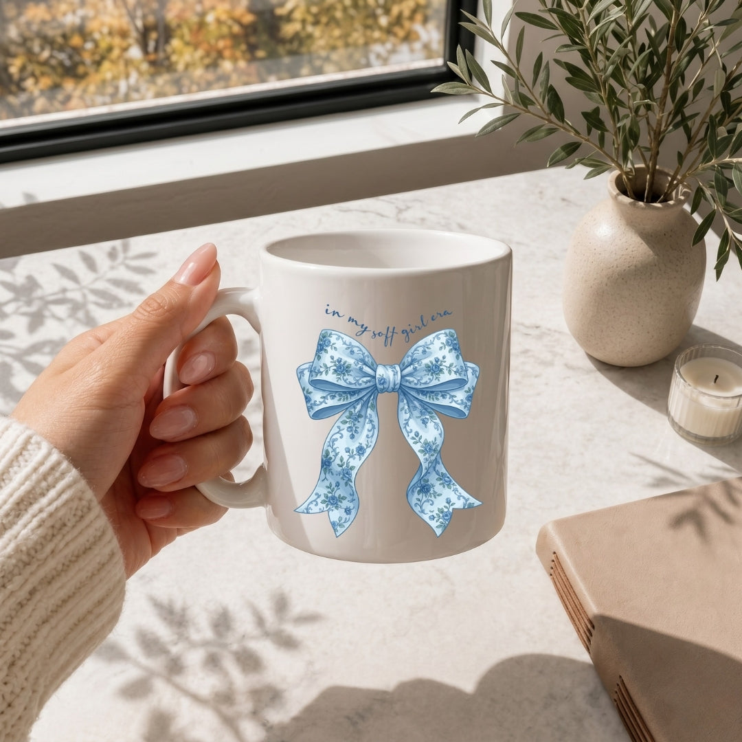 A woman holding a white ceramic mug featuring a blue floral bow graphic and the text "in my soft girl era."