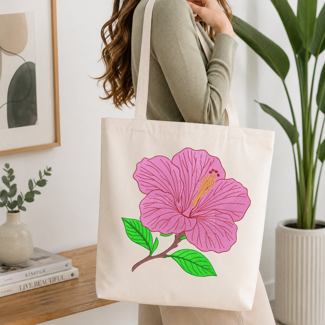 A woman wearing a sage green ribbed sweater carries a cream canvas tote bag with a large embroidered pink hibiscus flower and green leaves on the front, set in a bright, modern room with a wooden table and potted plants.