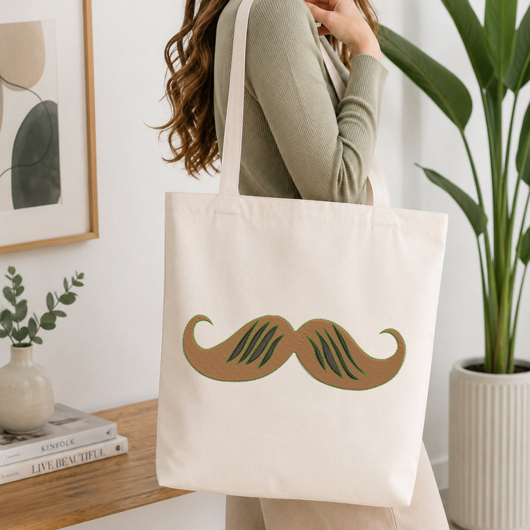 A person wearing a ribbed sage green top carries a cream canvas tote bag with a large brown and green mustache graphic on the front, set in a bright, modern room with a wooden table, books, and a potted plant.