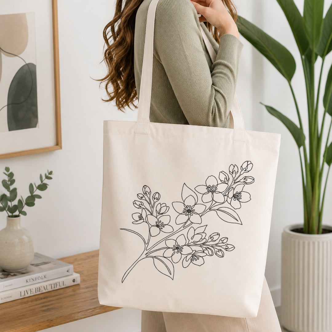 A woman wearing a sage green ribbed sweater carries a cream canvas tote bag with a black line art floral design. The scene is set in a bright, modern room with a wooden table, books, a small vase, and a large potted plant.
