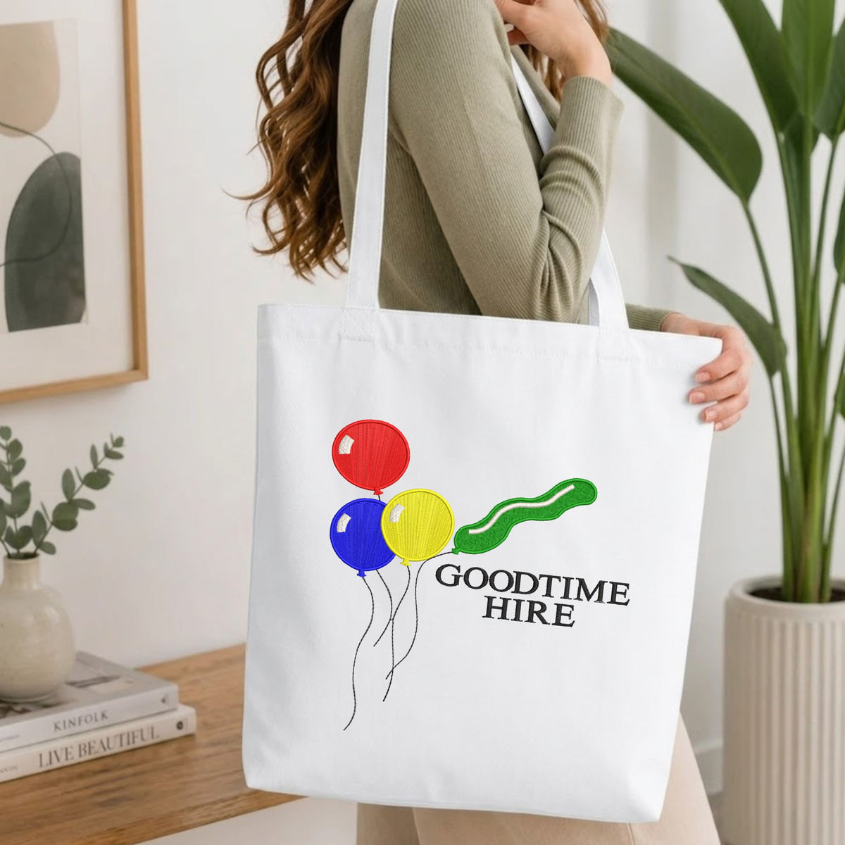 Woman carrying a white natural canvas tote bag featuring an embroidered colorful balloons and text design in a bright, modern interior setting