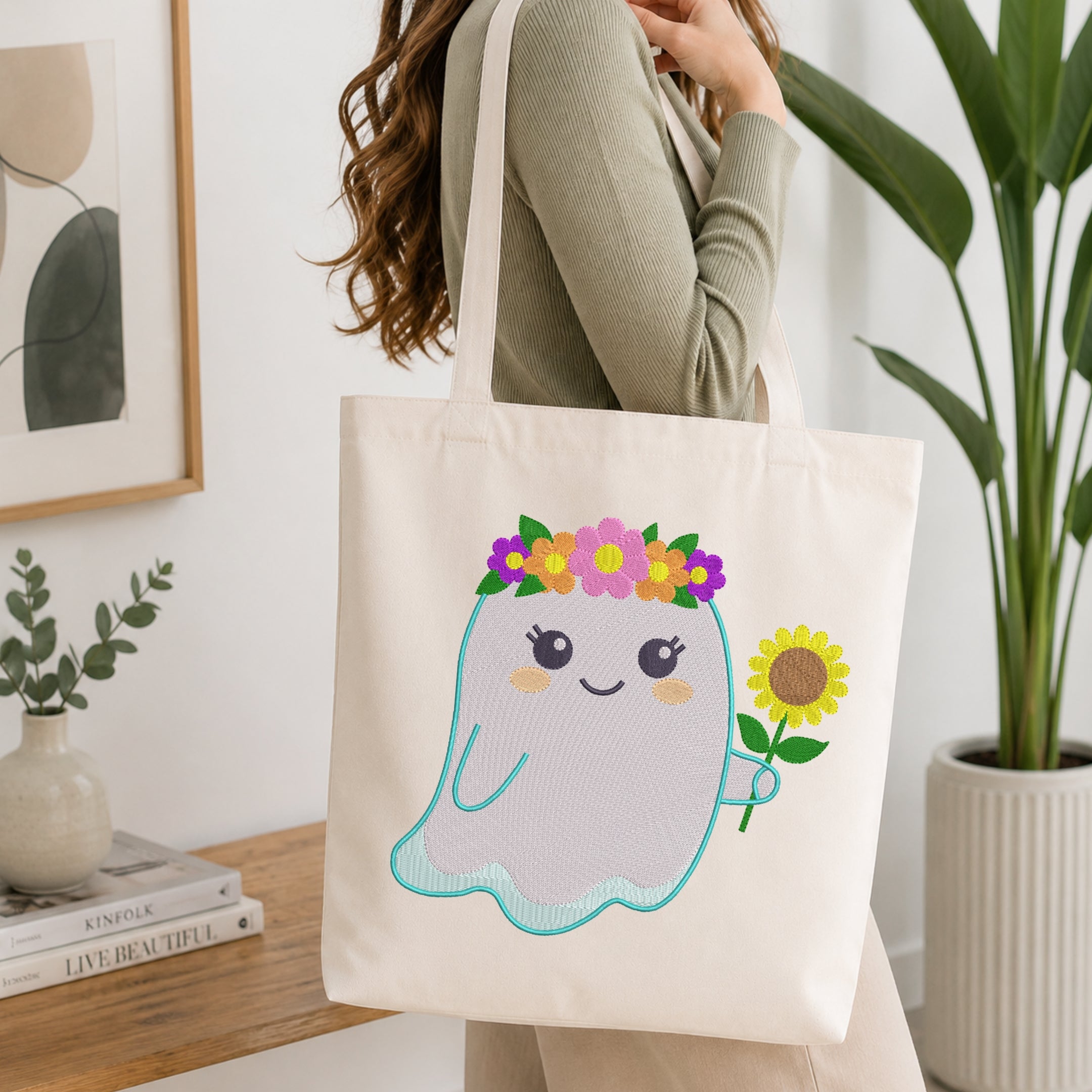 A woman carries a cream canvas tote bag with a cute cartoon ghost holding a sunflower and wearing a flower crown. The scene is set in a bright, modern room with a wooden table, books, and a potted plant.