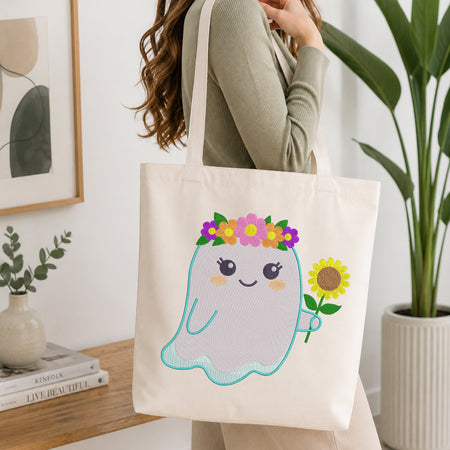 A woman carries a cream canvas tote bag with a cute cartoon ghost holding a sunflower and wearing a flower crown. The scene is set in a bright, modern room with a wooden table, books, and a potted plant.