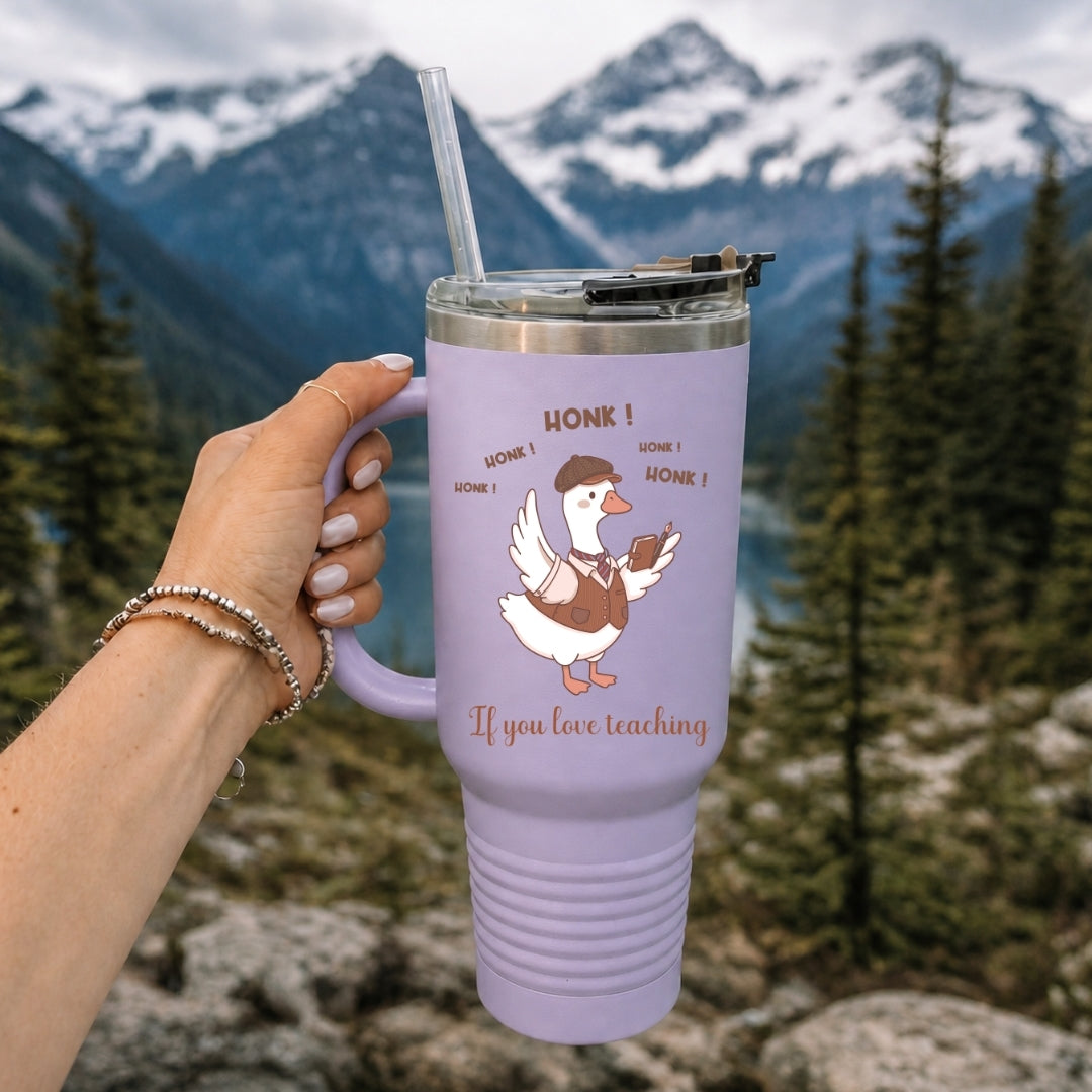 Hand holding a light purple insulated tumbler featuring the educator duck graphic, set outdoors against a scenic mountain and lake background.