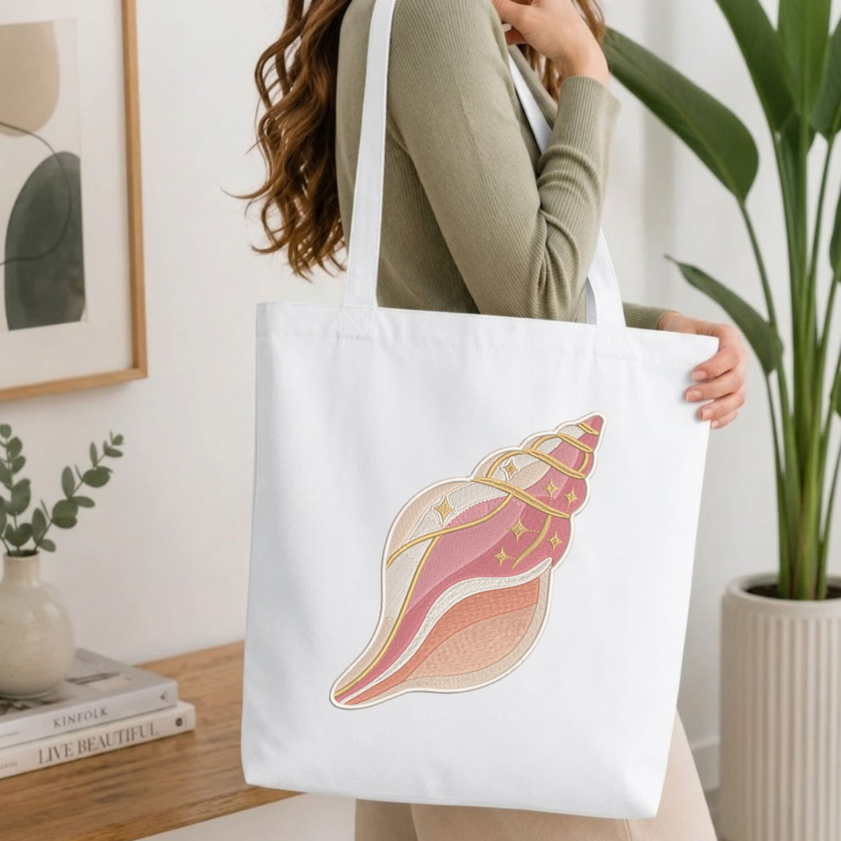 Woman carrying a white natural canvas tote bag featuring an embroidered pink and beige conch seashell design in a bright, modern interior setting.
