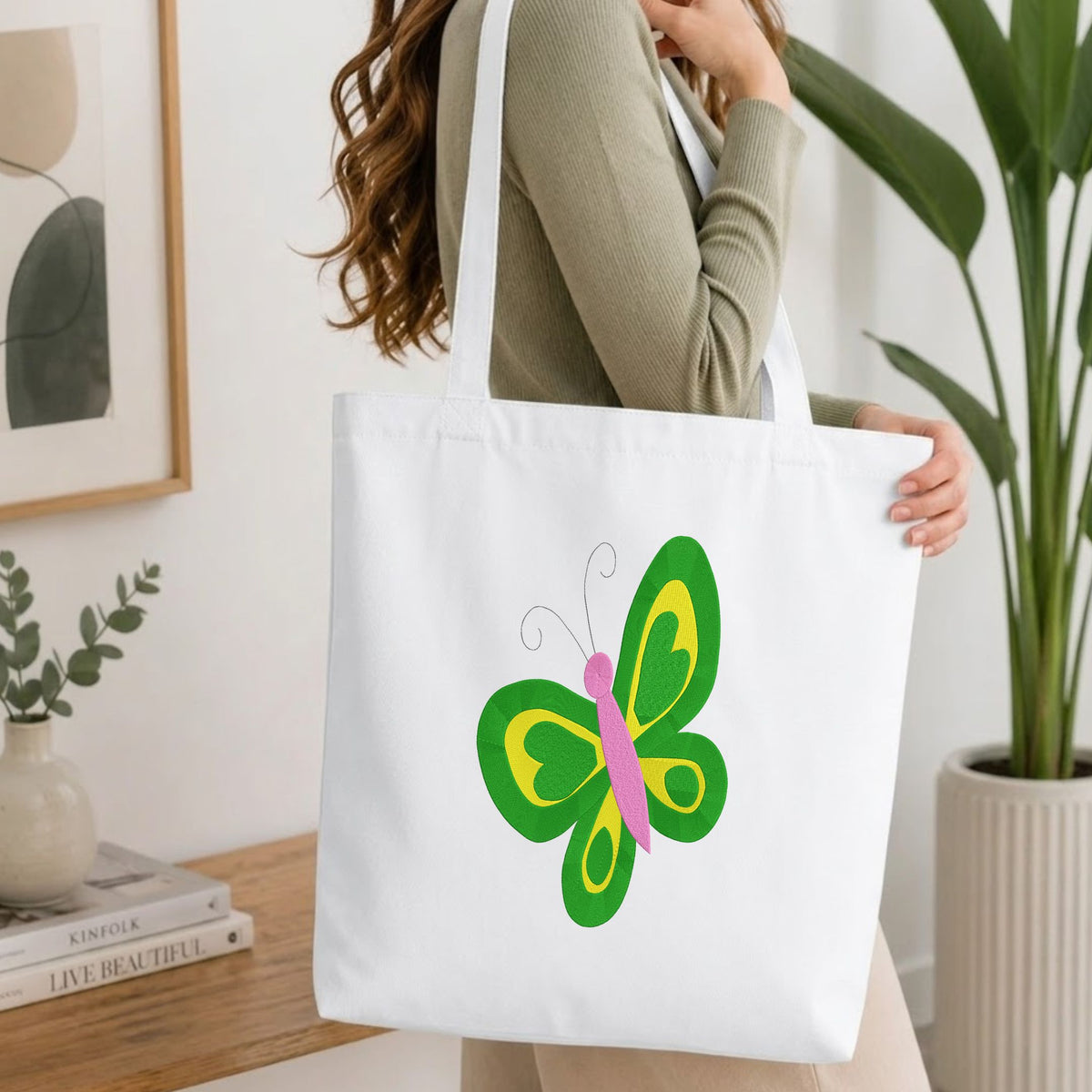 Woman carrying a white natural canvas tote bag featuring a green and yellow butterfly machine embroidery design in a bright, modern interior setting   