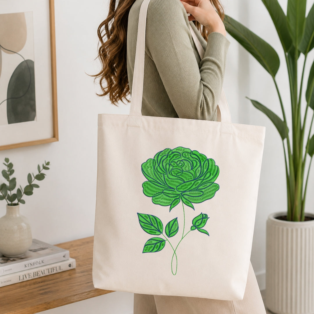 A woman wearing a ribbed sage green long-sleeve top carries a cream canvas tote bag with a large green rose illustration on the front, set in a bright, modern room with a wooden table, books, and a potted plant.